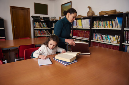 A mother helps her daughter with reading and writing at a library, surrounded by shelves of books...
