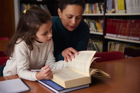 A woman guides a young girl through a book in a quiet library, fostering reading, learning, and...