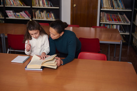 A caregiver guides a young girl as they read a large book in a warm school library, surrounded by...