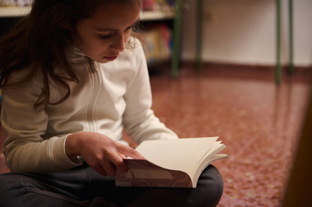 A child sits on the library floor, turning pages with concentration, surrounded by a calm, studious atmosphere.の写真素材