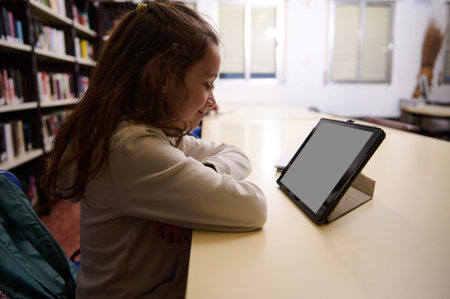A young student sits at a library table, using a tablet for study. Bright shelves and a quiet atmosphere evoke learning, reading, and homework in a school library.の写真素材
