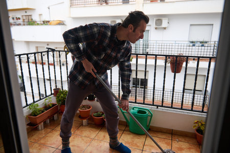 A man on an urban apartment balcony mops the tiled floor while surrounded by potted plants, a green watering can sits nearby, creating a lived-in, domestic scene.の写真素材