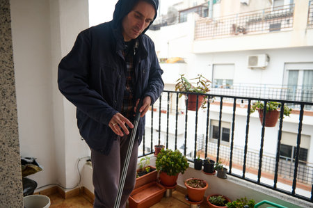 A senior man in a hooded jacket on a city balcony adjusts his cane while tending potted plants: a...