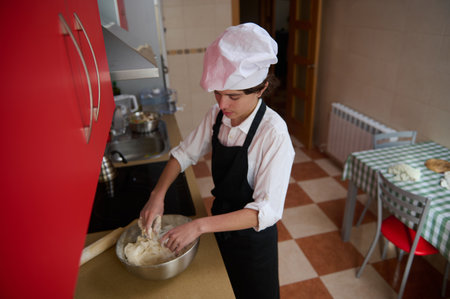 A young cook in a white chef hat and black apron mixes dough in a small kitchen. Red refrigerator,...