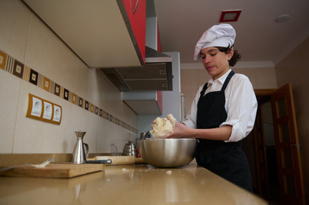 A young cook in a white uniform and chef hat kneads dough by hand in a modern kitchen, using a...