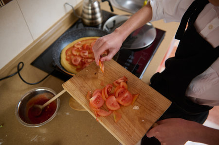 A cook in an apron prepares fresh tomato slices on a wooden cutting board for a homemade pizza. Tomato sauce simmers in a pan nearby, as the kitchen buzzes with cooking activity.の写真素材