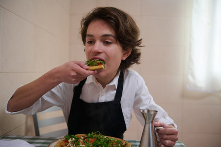 A young man in a white shirt and black apron enjoys a fresh vegetable-topped pizza in a bright, homey kitchen.の写真素材