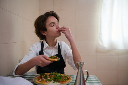A young person in a white shirt and black apron bites a slice of pizza at a checkered table, with a ready pie and a metal bottle nearby in a simple kitchen.の写真素材