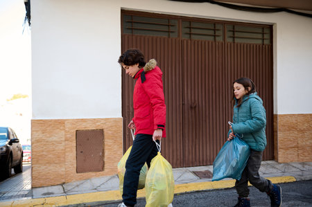 Two kids in winter jackets carry plastic bags along a sidewalk near a building with brown doors. A candid street scene capturing everyday shopping, family outing, and urban life.の写真素材