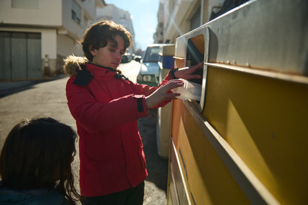 A young boy in a red winter jacket posts a letter into a roadside mailbox while a girl looks on. Warm sunlight and urban surroundings evoke everyday family moments.の写真素材