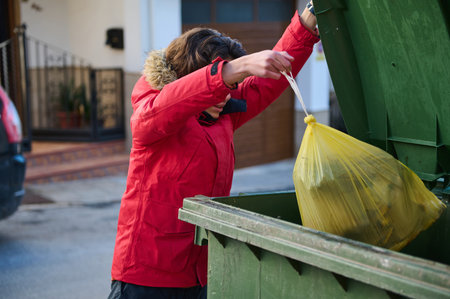 A person in a red jacket deposits a yellow trash bag into a green dumpster on an urban street,...