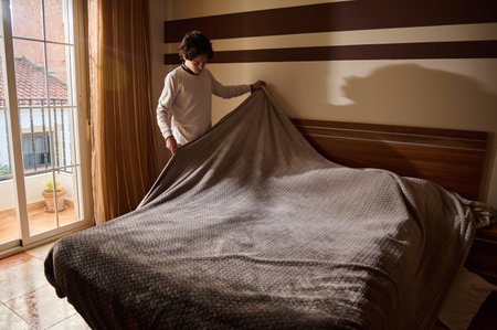 A person folds back a warm blanket to tidy a home bedroom, inviting sunlight through a balcony door....