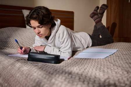 A student lies on a bed, focused on writing in a notebook. A pencil case and spiral notebook lie...