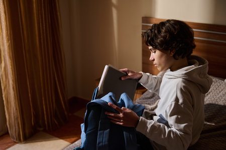 Young student sits on a bed in a cozy room, opening a notebook and preparing a backpack for study....