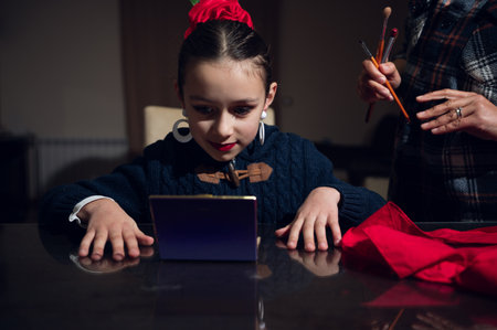 A young girl with a red bow sits at a reflective table as brushes and makeup tools are nearby, capturing a warm, creative moment of learning and style.の写真素材