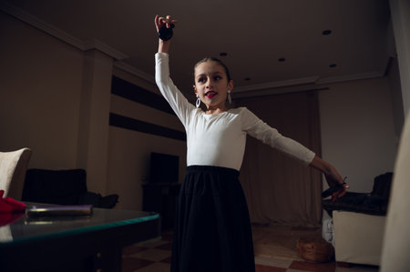 A young girl dancer stands in a cozy living room, wearing a white top and black skirt, with one arm raised, showcasing confidence, grace, and focus.の写真素材