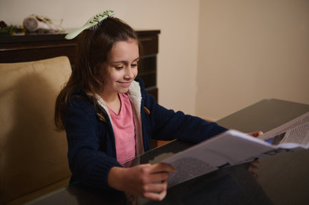 A smiling young girl sits at a glass dining table reading a magazine. She wears a pink shirt and...