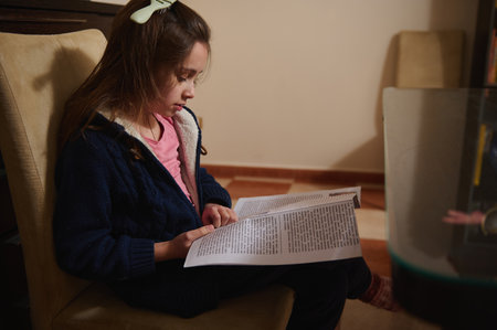A young girl in a navy cardigan and pink shirt reads a book while seated comfortably at home. Warm light conveys a calm, focused moment of learning and family life.の写真素材