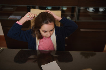 A young girl sits at a kitchen table, looking down in concentration. Casual home setting with a pink shirt and navy cardigan, a sheet of paper on the table.の写真素材