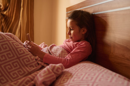 A young girl rests in bed under a pink blanket, scrolling on her phone in warm, cozy lighting. A...