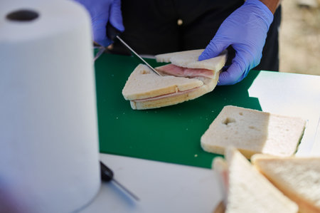 A person in blue gloves slices ham between bread on a green cutting board. Extra sandwich slices sit...