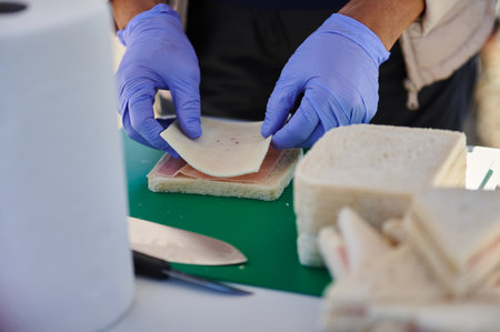 A food preparer wearing blue gloves layers ham and cheese on white bread at a deli station. Close-up...