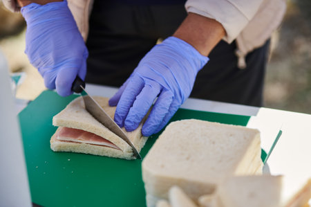 A cook wearing purple gloves slices a ham sandwich on a green cutting board, with bread nearby,...
