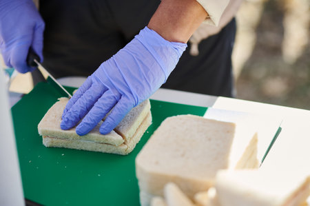 Gloved hands slice and assemble a sandwich on a green cutting board, showcasing food safety in...