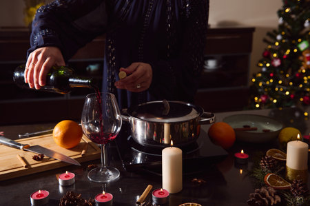 A stylish woman in a dark gown pours red wine into a glass as candles glow beside a simmering pot, oranges, and a sparkling Christmas tree for a cozy holiday kitchen setting.の写真素材