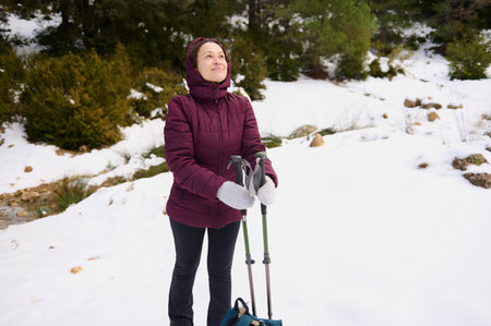 A woman in a burgundy jacket stands with trekking poles in a snowy landscape, enjoying a calm moment in nature during a winter hike.の写真素材