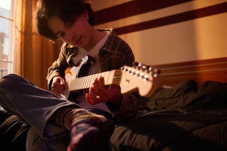 A young man sits on a bed, playing an electric guitar as warm sunlight fills a cozy room, capturing...