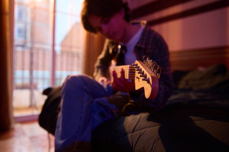 A musician sits on a bed, playing an electric guitar in a softly lit, intimate room. The warm glow...