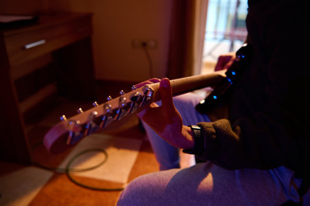 A focused musician tunes an electric guitar in a cozy indoor space. The warm lighting highlights the...