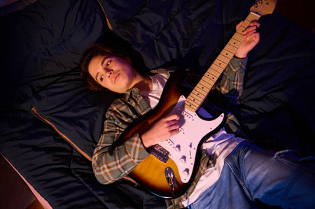 A young musician relaxes on a dark quilt and plays a sunburst electric guitar. Casual plaid shirt, blue jeans, and moody lighting create a warm, intimate home music moment.の写真素材