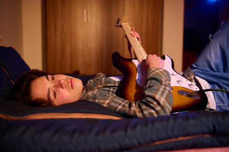 A young woman lies on a bed, playing a sunburst electric guitar. Casual, cozy scene conveying creativity, relaxation, and musical joy in a bedroom.の写真素材