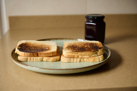 A simple breakfast scene featuring two lightly charred toast slices on a plate with a jar of jam in...