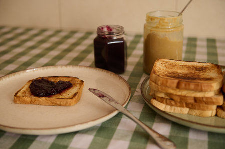 A cozy kitchen scene featuring a slice of toasted bread with jam, a jar of jam, and a peanut butter jar beside a stack of toast on a green checkered tablecloth.の写真素材