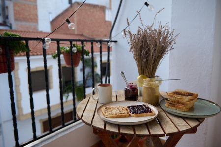 A sunny balcony breakfast scene with toast, jam, a mug, condiments, and dried flowers on a wooden table, offering a warm, intimate outdoor meal mood.の写真素材