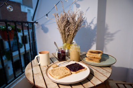 A warm balcony breakfast setup with toasted bread spread, jam and peanut butter, a mug, jars, and a dried wheat vase under string lights for a cozy morning mood.の写真素材