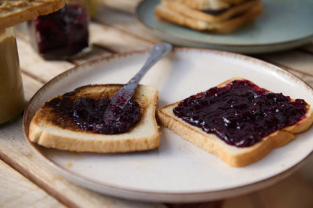 Two slices of toasted bread spread with dark berry jam on a plate, with a rustic wooden table and...