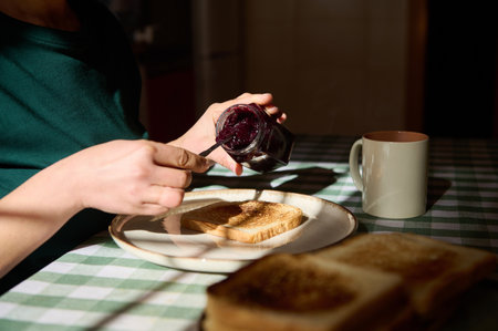 A person spreads dark berry jam on toast at a sunny breakfast table, with a mug nearby, capturing a...