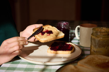 A cozy breakfast scene: toast with peanut butter and jam being spread by hands at a checkered table....