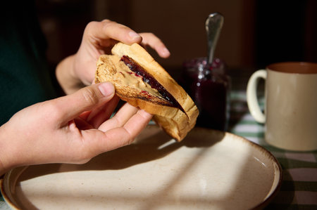 Close-up of hands holding a peanut butter and jelly sandwich over a plate, with jam jar and mug in...