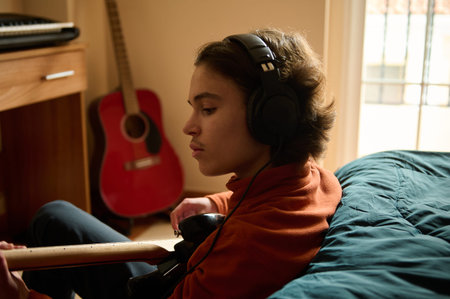 A teen boy in an orange hoodie sits on a bed, wearing headphones, playing a guitar in a sunlit...