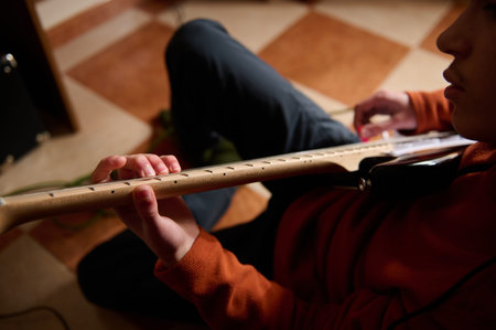 A teen of Middle Eastern ethnicity plays an electric guitar indoors, hands on the neck, showing...