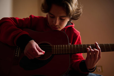 A teen boy in a red hoodie concentrates on strumming an acoustic guitar in a warm, indoor space,...