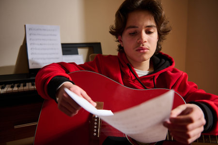 A teen boy of Middle Eastern ethnicity tunes a red guitar while reading sheet music, showing focus...