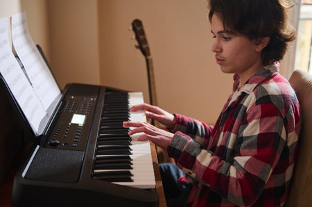 A teen boy in a plaid shirt plays a keyboard, focused on the music. Sheet music rests on the stand while a guitar sits in the background.の写真素材