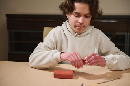 A teen boy sits at a light table, shaping red clay with his hands. He appears calm and focused, enjoying a simple home hobby project.の写真素材