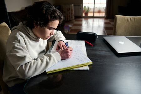 Teenage boy focused on homework at a cozy home table in the evening studying with notebook pen...